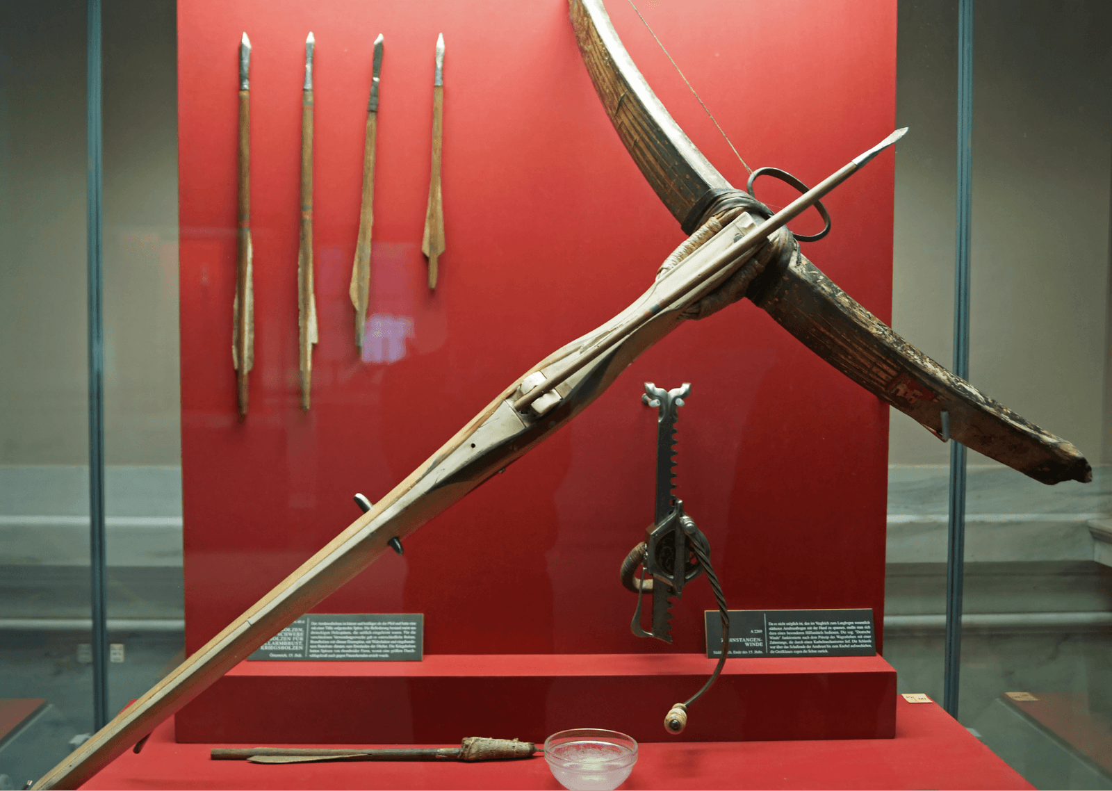 A historical museum display featuring an authentic medieval crossbow loaded with a bolt, alongside additional arrows and a cranequin spanning mechanism.