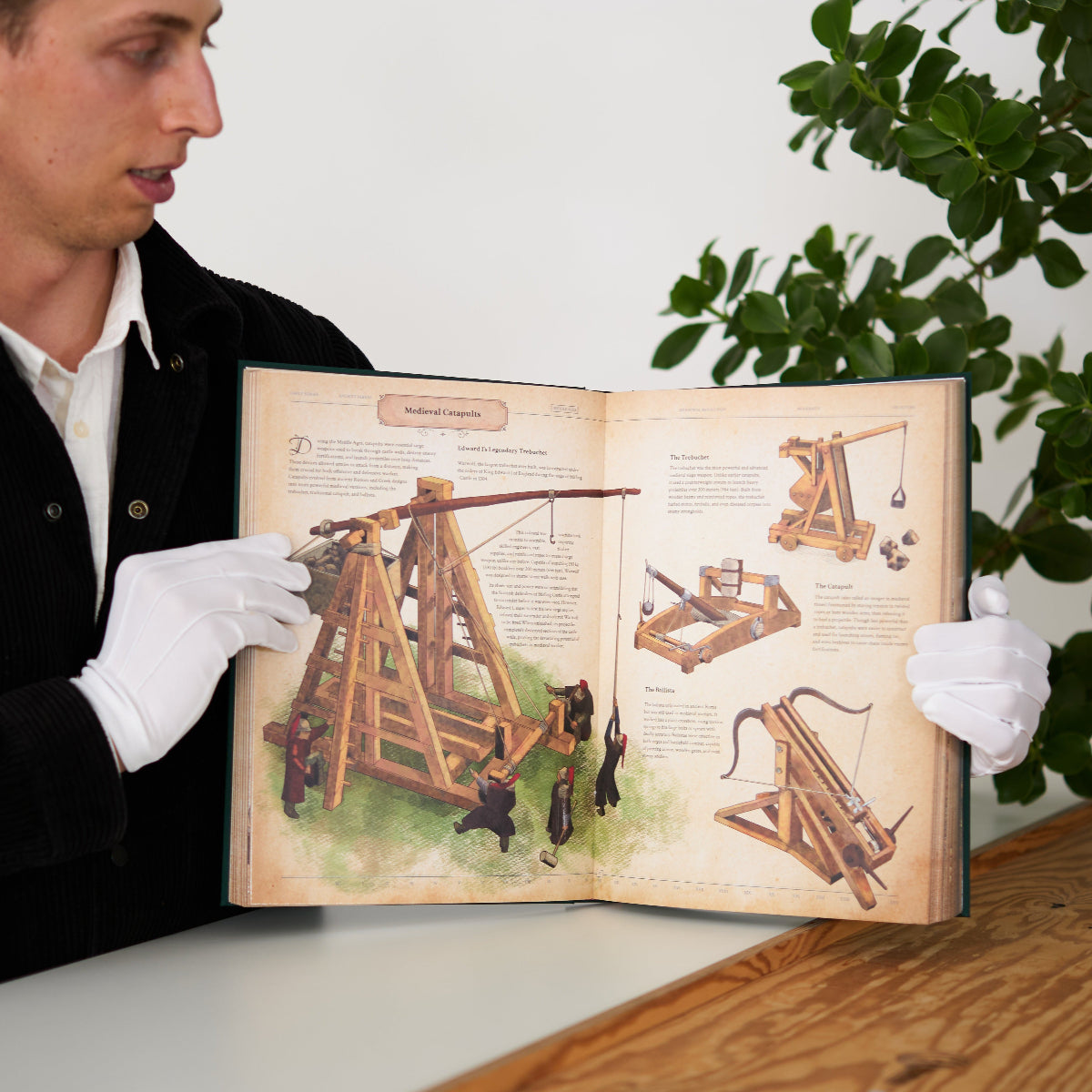 Person holding an open book with illustrations of mechanical devices, surrounded by plants.