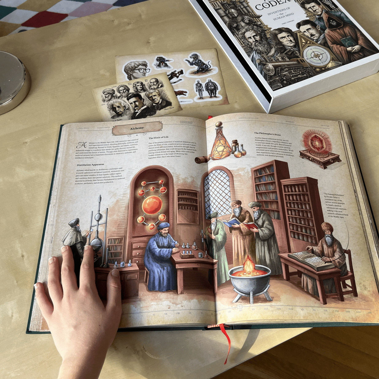 The Codex book open to the Alchemy section alongside its illustrated slipcase, sticker sheet, and art card.
