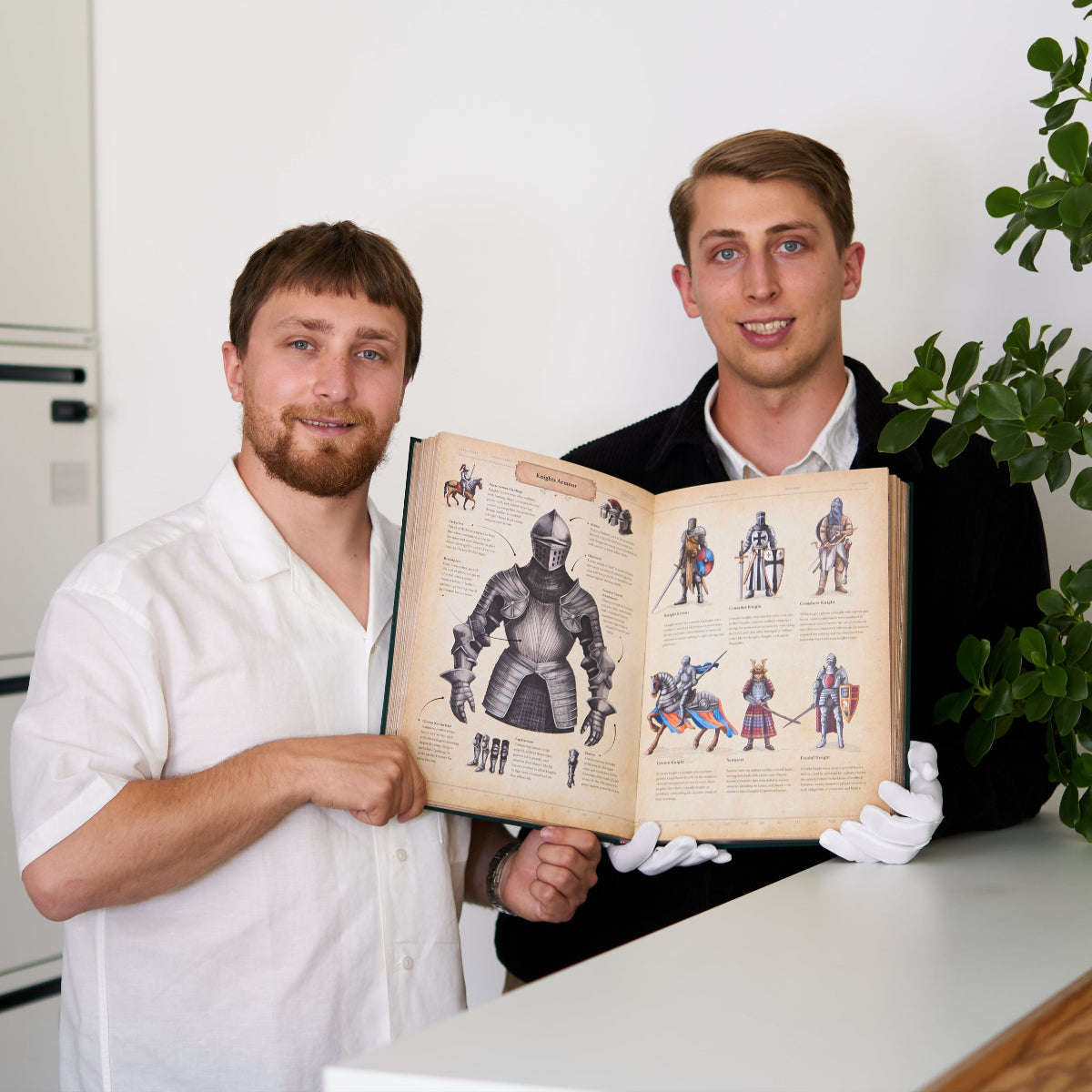 Two men holding The Codex open to a spread on medieval knight armor, showing detailed illustrations of suits of armor, weapons, and warriors.