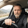 Close-up portrait of a middle-aged man with a grey beard sitting at the steering wheel of a car.