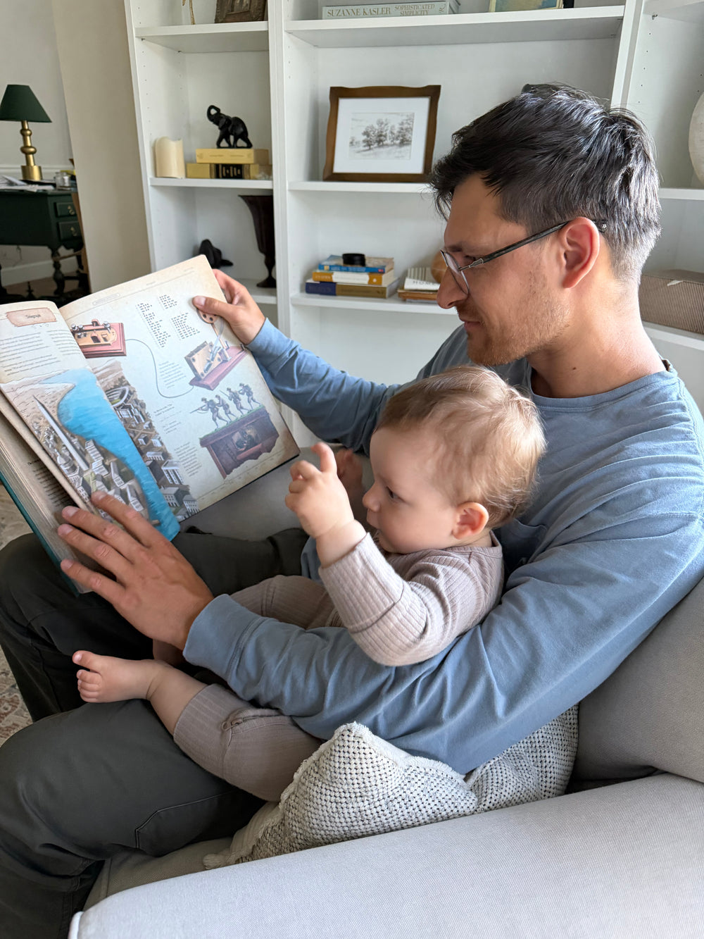 Man with glasses sitting on an armchair reading an open hardcover history book to a toddler.