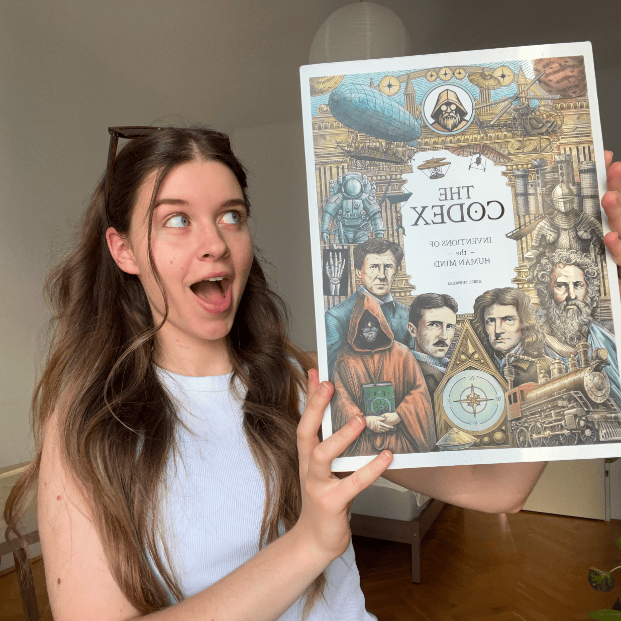 An excited young woman holding up the illustrated slipcover for The Codex Inventions of the Human Mind.