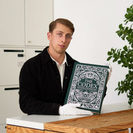 Man holding The Codex Book of inventions , a large illustrated kids encyclopedia of inventions in Da Vinci-inspired style, showing its size and ornate green cover.
