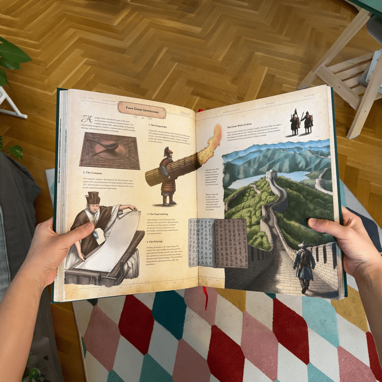 Hands holding open The Codex book to a spread featuring gunpowder, paper making, and a panoramic Great Wall illustration.