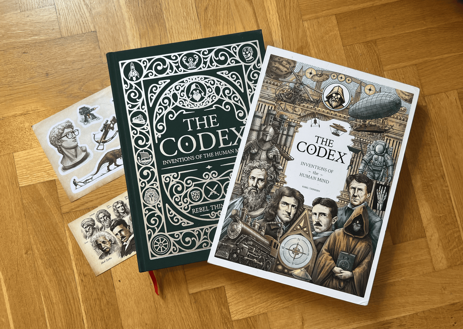 Flat lay of The Codex Inventions green hardcover, illustrated slipcase, sticker sheet, and art card on wood floor.