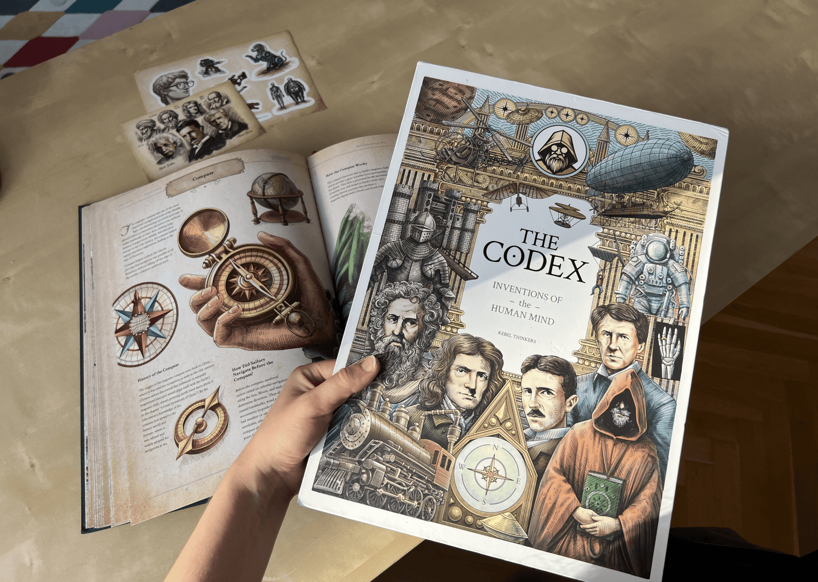 he Codex slipcover held above open pages showing compass illustrations, stickers, and a portrait card.