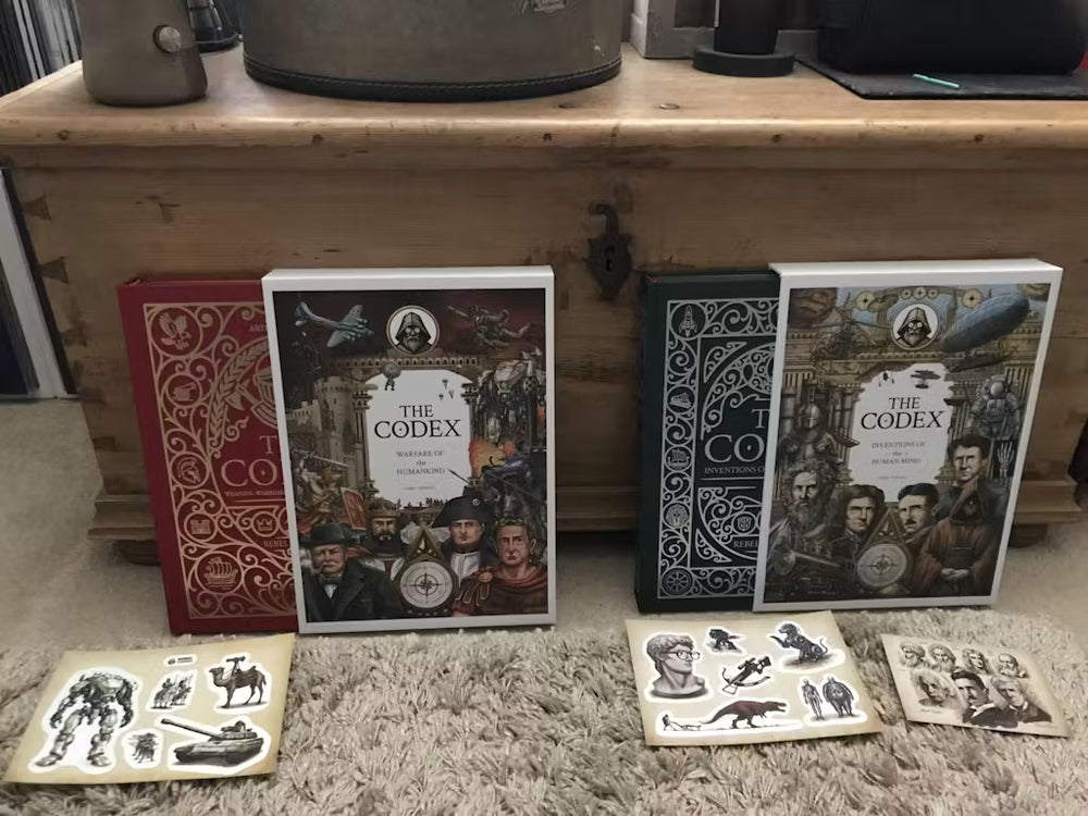 Red and green copies of The Codex books with illustrated slipcases and bonus sticker sheets on a carpet.